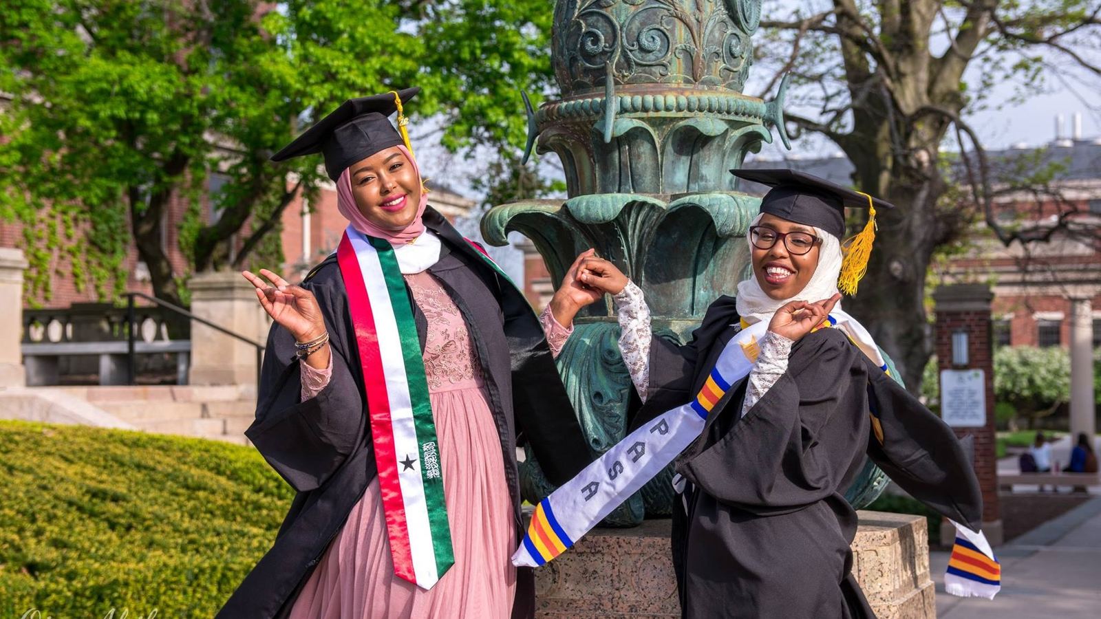 Two Abaarso graduates in caps and gowns smiling at their college commencement, one wearing a Somaliland flag stole.