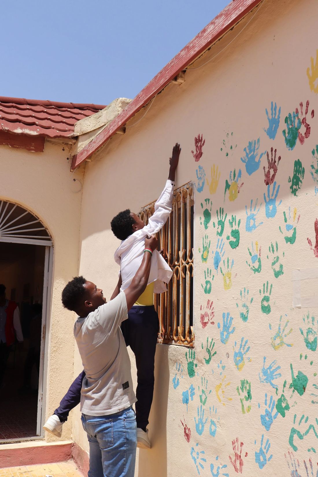 A young man lifts a boy so he can press his handprint high on a school wall covered in painted handprints.